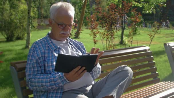 Um homem idoso com óculos lendo um e-book em um banco de estacionamento — Vídeo de Stock