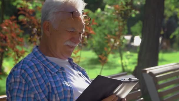 Vista lateral de un anciano con bigote está leyendo momento divertido en el libro. El viejo está sentado en el banco en el parque y leyendo ebook — Vídeo de stock
