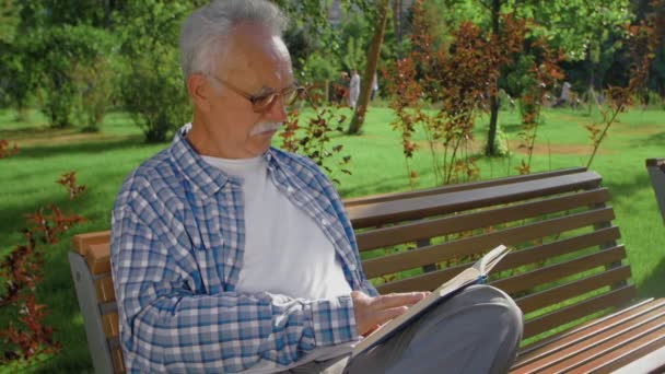 Ein älterer Mann sitzt auf der Bank und liest Buch im Park — Stockvideo