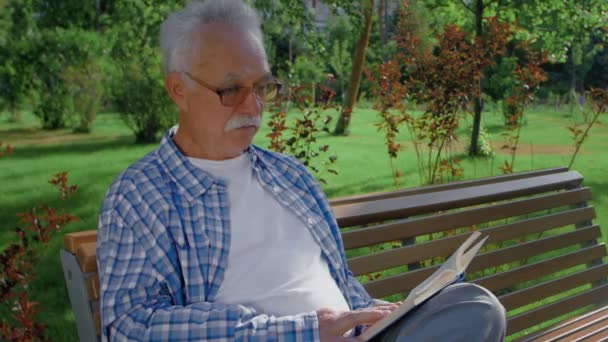 Un hombre mayor sentado en el banco y leyendo un divertido libro en el parque . — Vídeo de stock