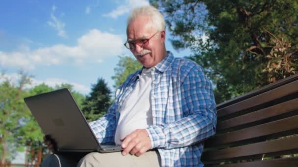 Glad senior man talar i skype, sitter på bänken i trädgården. En pensionär har en laptop i knät. Han viftar med handen för att säga adjö — Stockvideo
