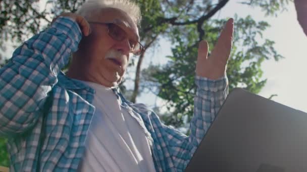 Gamle man i glasögon försöker använda datorn i parken, men han kan inte. Han ringer nån som kan hjälpa honom. — Stockvideo