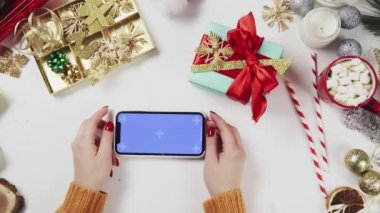 Chroma anahtarı olan akıllı telefon kullanan bir kadın, tıklatıyor, kaydırıyor, kaydırıyor. Beyaz masa arkasında Noel tatili süslemesi.