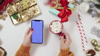 Kakao içen ve Chroma anahtarıyla akıllı telefon kullanan bir kadın. Beyaz masa arkasında Noel tatili süslemesi.