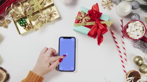 クロマキーでスマートフォンを使用して女性,タップ,スワイプ,上にスクロール.白いテーブルの背景にクリスマスの休日の装飾. — ストック動画