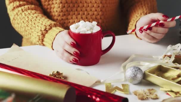 Frau im orangefarbenen Pullover trinkt heiße Schokolade mit Weihnachtsgeschenken auf Tisch — Stockvideo