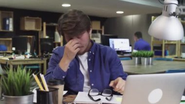 Ofis masasında dizüstü bilgisayar başında oturup kahve içmekten yorgun düşmüş genç bir adam.