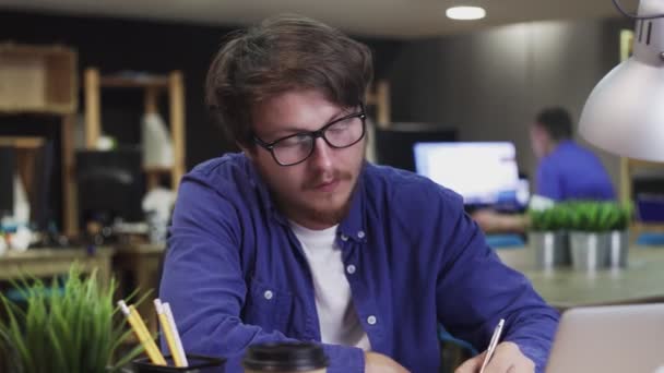 Молодой человек в очках что-то пишет в блокноте в офисе — стоковое видео