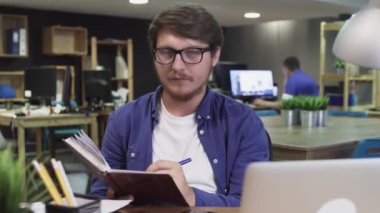 Elinde not defteri olan çekici bir adam modern ofiste kameraya bakıyor.