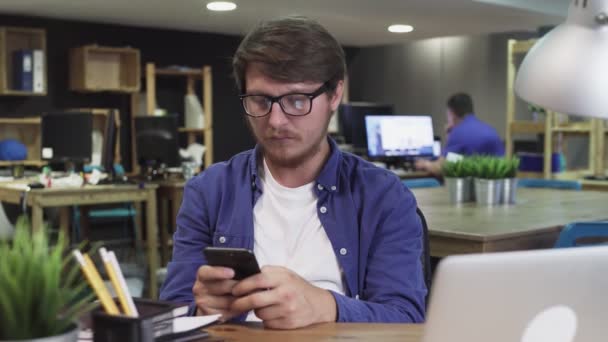 Jeune homme utilise Smartphone alors qu'il est assis au bureau. Homme navigue sur Internet, Montres Vidéos et utilisations Réseaux sociaux — Video