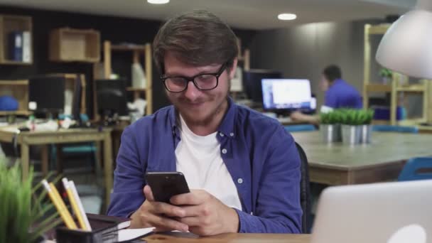 Happy Young Man utilise Smartphone alors qu'il est assis au bureau. Homme navigue sur Internet, Montres Vidéos et utilisations Réseaux sociaux — Video