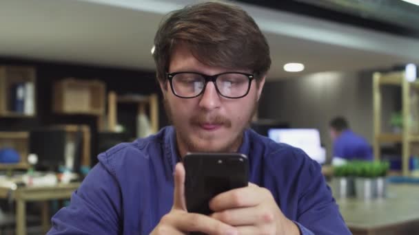 Портрет счастливого молодого человека использует смартфон во время работы в офисе. Человек просматривает через Интернет, смотрит видео и использует социальные сети — стоковое видео