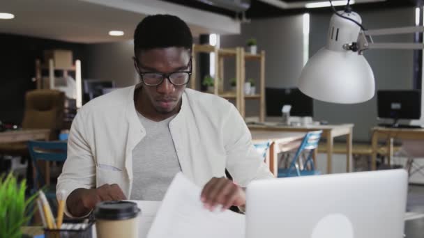 若いですよく見るアフリカ人男性で眼鏡をかけてオフィスのテーブルに座って書類を扱う — ストック動画