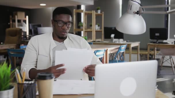 Ung afrikansk man i glasögon sitter vid kontorsbordet och arbetar med dokument — Stockvideo