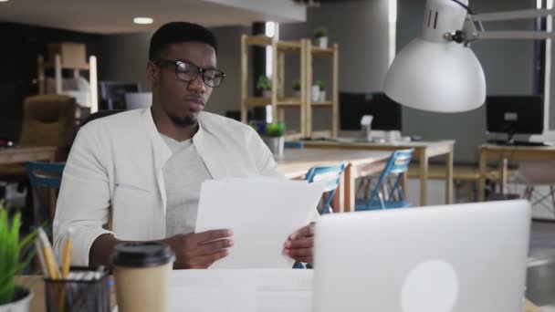 Молодой африканский мужчина в очках сидит за рабочим столом и читает документы — стоковое видео