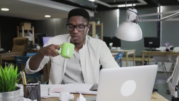 Giovane uomo africano con le cuffie sta lavorando su laptop e bere caffè o tè in ufficio — Video Stock