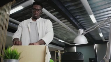 Afrikalı adam yeni ofise taşınıyor. Kişisel eşyalarını karton kutuya koyuyor.