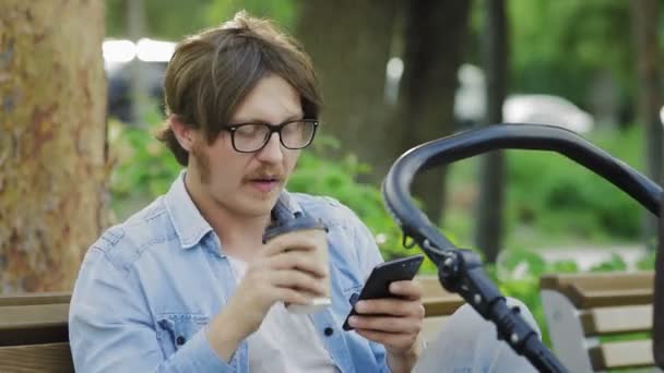 O bebê está dormindo em um carrinho, o pai está usando um celular e bebendo café . — Vídeo de Stock