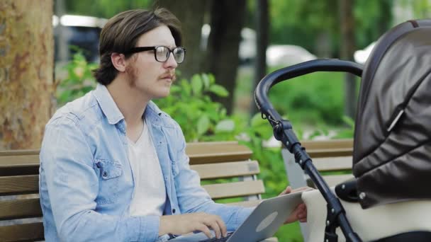 Ung man i glasögon arbetar bärbar dator och ler till spädbarn i vagn, multitasking, frilans — Stockvideo