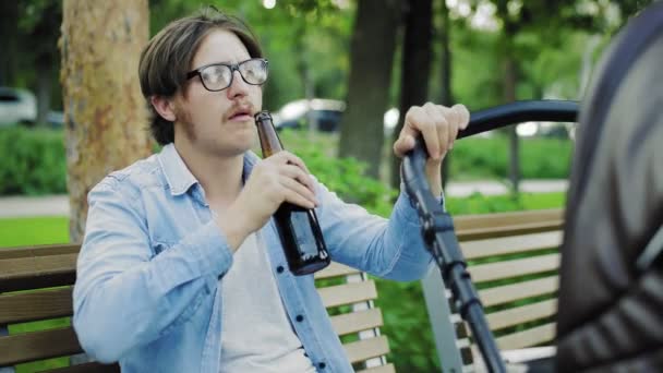 Alcolismo in un luogo pubblico. Un uomo ubriaco che beve birra e dondola passeggino in un parco — Video Stock