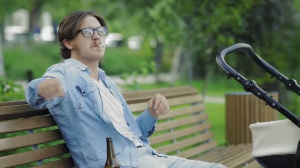 Молодой отец курит и пьет пиво, размахивая коляской в парке — стоковое видео