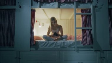 Genç ve çekici bir kadın yurt odasında yatakta oturuyor ve gece akıllı telefon kullanıyor.