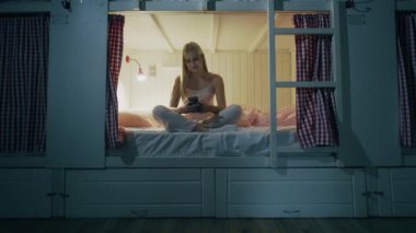 Mutlu, gülümseyen bir kadın yurt odasında yatakta oturuyor ve gece akıllı telefon kullanıyor.