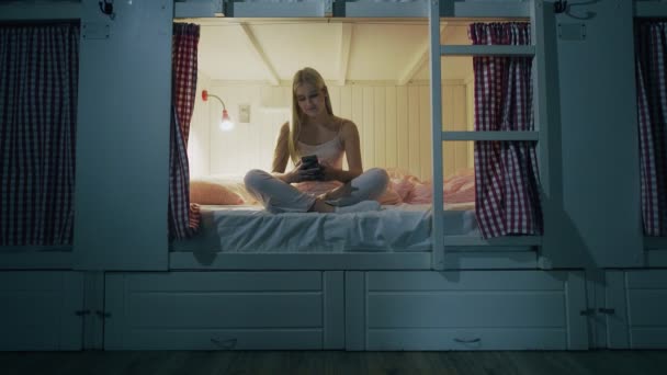 Mujer sonriente feliz sentado en la cama en la habitación del albergue y el uso de teléfono inteligente por la noche — Vídeo de stock