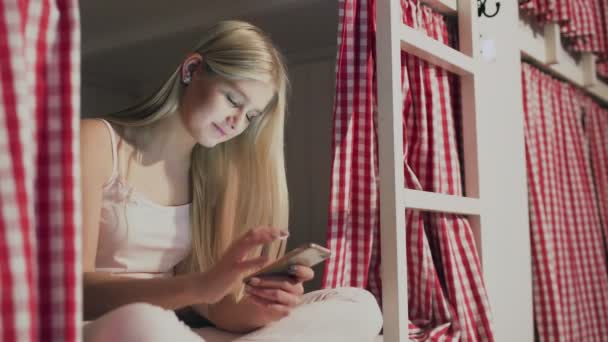 Young attractive woman sitting on the bed in the hostel room and using smartphone — Stock Video