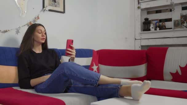 Mujer atractiva joven sentado en el sofá y el uso de teléfono inteligente que tiene chat de vídeo — Vídeos de Stock