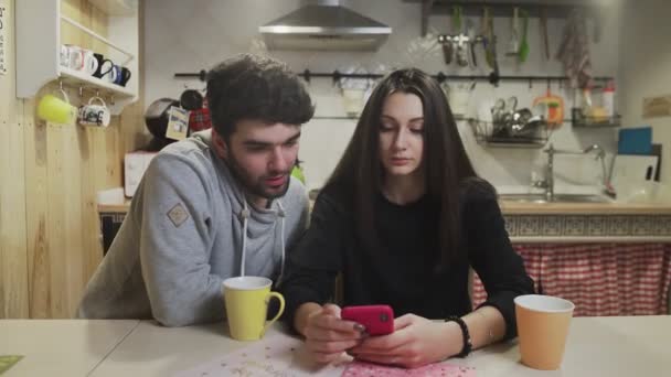 Genç çift mutfakta kahvaltı yapıyor. Akıllı telefon ekranını izleyen neşeli bir çift. Mutfakta kadınla konuşan mutlu bir adam.. — Stok video