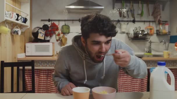 Um jovem a tomar o pequeno-almoço na cozinha. Homem come flocos de milho Cereais e bebe um café — Vídeo de Stock