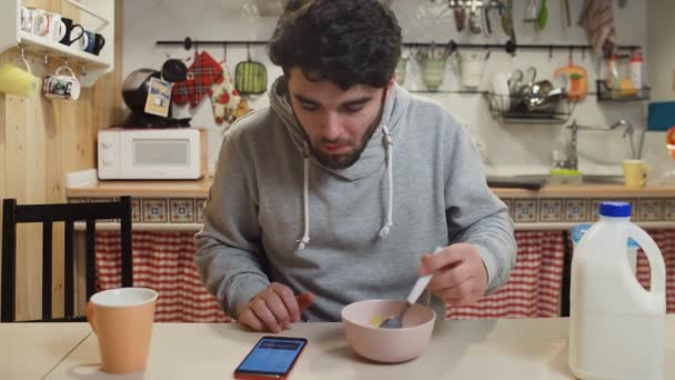 Mladý muž se snídaní v kuchyni. Člověk jí kukuřičné vločky obiloviny a používá smartphone — Stock video