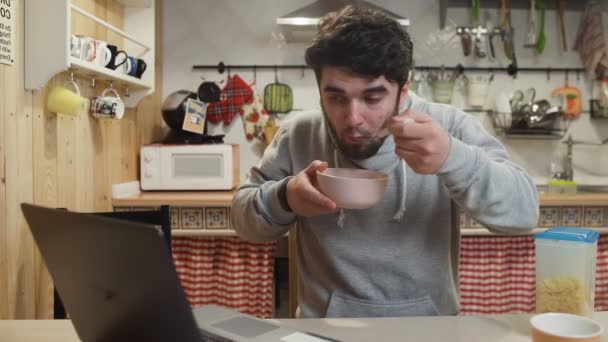 Mladý muž pracuje notebook a jí kukuřičné vločky obiloviny doma kuchyně. — Stock video