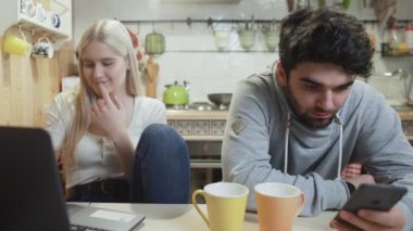 Genç çift konuşuyor ve mutfakta modern bilgisayar ve telefon kullanıyor..