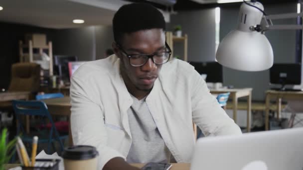 Portre genç Afrikalı Amerikalı adam dizüstü bilgisayarda çalışıyor ve ofiste telefon kullanıyor. — Stok video