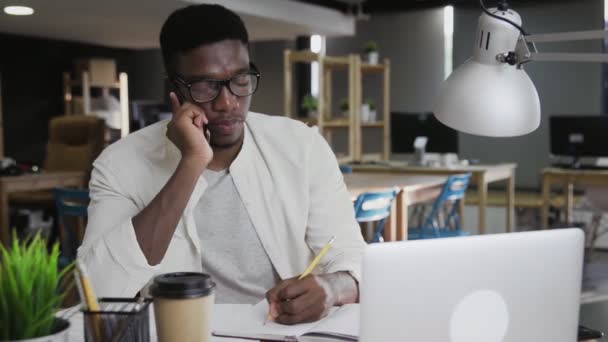 Hombre africano está hablando en el teléfono móvil y tomando nota en la oficina moderna — Vídeos de Stock