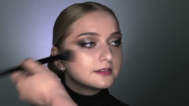 Макияж художник делает профессиональный макияж для молодой женщины в студии красоты. Макияж художник наносит румяна большой кистью — стоковое видео