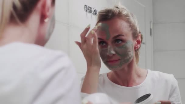 Woman applying clay mask on her face looking in mirror — Stock Video