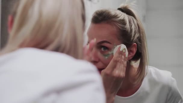 Woman removes clay mask on her face looking in mirror — Stock Video