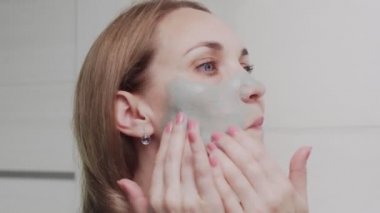 Yetişkin bir kadının aynaya bakarak yüzüne kilden maske takması.
