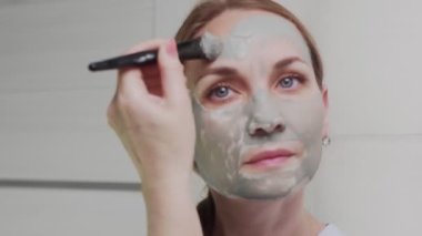 Yetişkin bir kadının kameraya bakarak yüzüne kilden maske takması.
