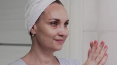 Kafasında havlu olan yetişkin bir kadın losyon ya da yağ sürüyor ve aynaya bakıyor.