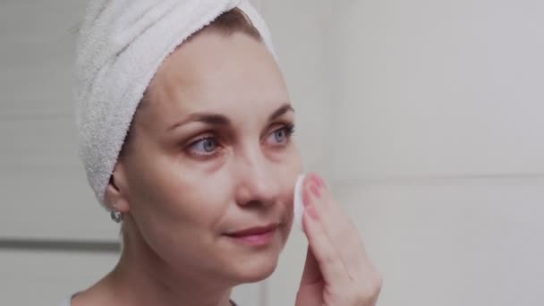 Volwassen vrouw met een handdoek op haar hoofd aanbrengen lotion en kijken in spiegel genieten van natuurlijke schoonheid en gezond — Stockvideo