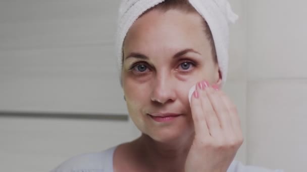 Доросла жінка з рушником на голові наносить крем і дивиться на камеру — стокове відео