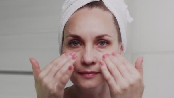 Adult woman with a towel on her head applying cream and looking at the camera — Stock Video