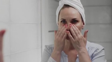 Kafasında havluyla hayal kırıklığına uğramış yetişkin kadın banyo aynasında yüzüne bakıyor.