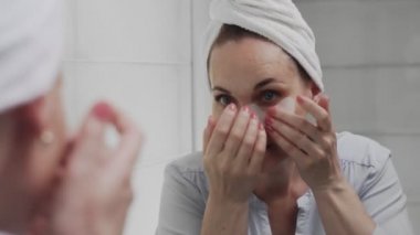 Kafasında havlu olan yetişkin bir kadın gözlerinin altına kolajen pedleri koyar ve aynaya bakar.