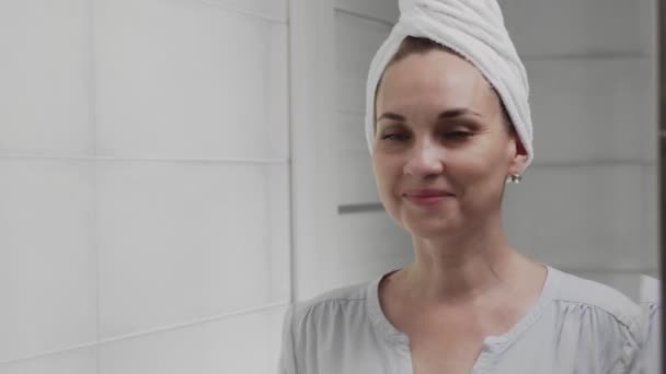 Λευκή γυναίκα με μια πετσέτα στο κεφάλι μπροστά από έναν καθρέφτη στο μπάνιο. Πλύσιμο προσώπου, περιποίηση δέρματος, ευεξία — Αρχείο Βίντεο