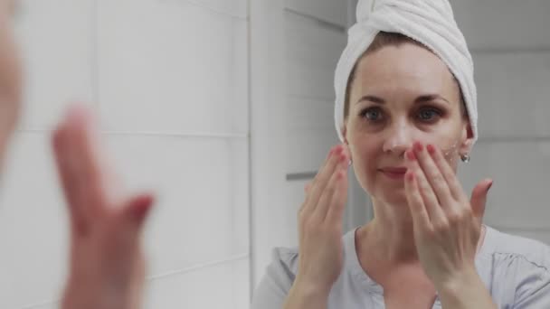 Доросла жінка з рушником на голові наносить крем і дивиться у дзеркало насолоджується природною красою і здоровим — стокове відео